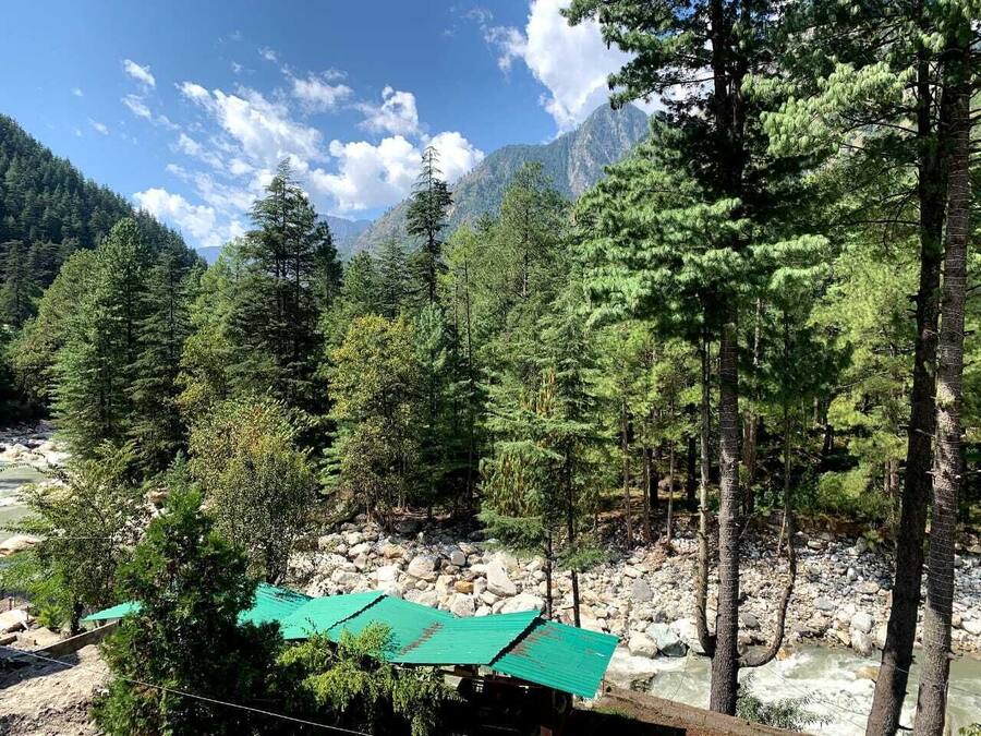 Un paisaje montañoso con un río, un bosque denso y un edificio pequeño con techo verde.