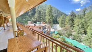A wooden balcony with a view of a river and forested mountains. - Hotel Purnima River View (Bhuntar)
