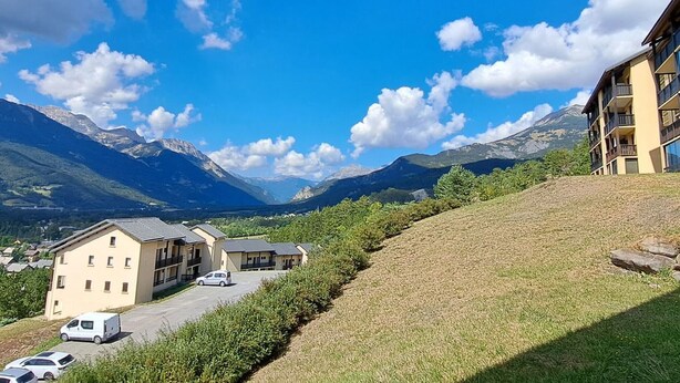 A residential area with a building featuring a parking lot and a hillside with a view of mountains. - Cozy Mini T2 in Barcelonnette - For 4, sunny balcony (Barcelonnette)