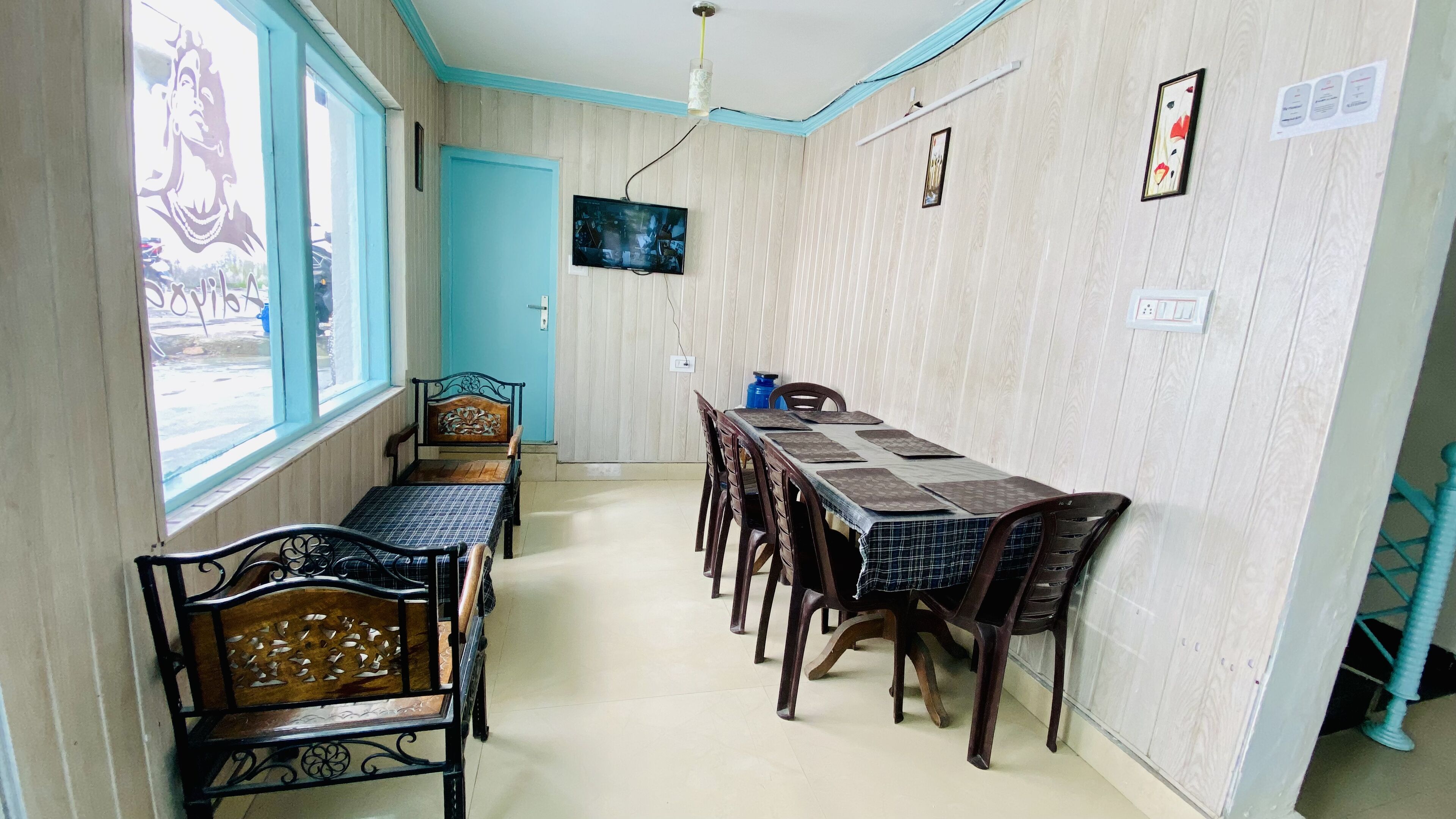 A dining area with a long table, several chairs, a television mounted on the wall, and a window with a view of the outside.