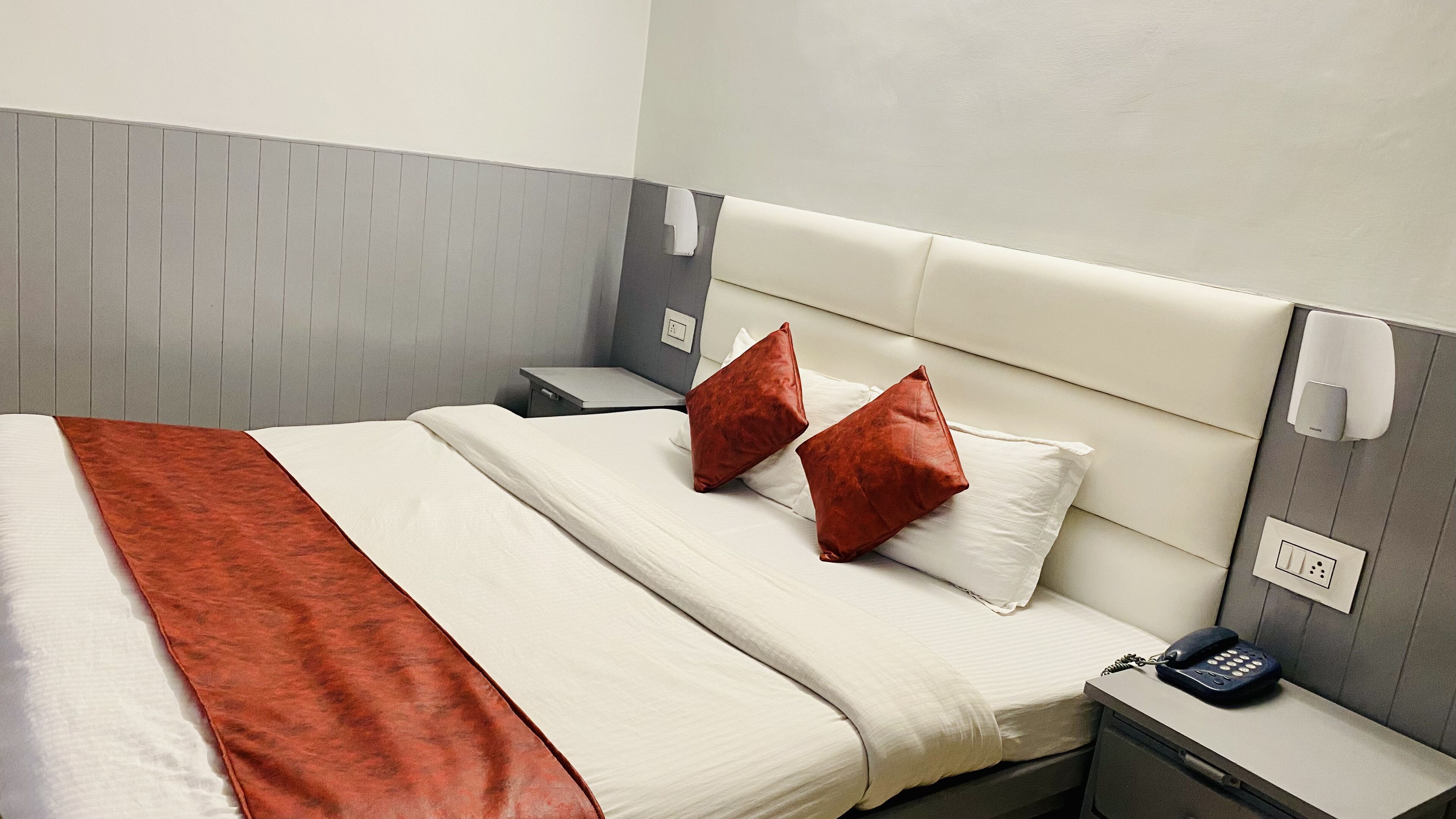 A neatly made bed with a white and red bedspread, two red pillows, a bedside table with a phone, and a wall-mounted light.