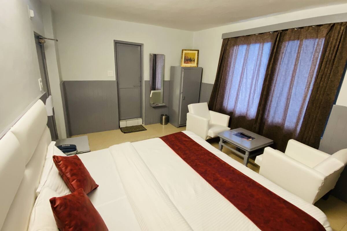 Hotellihuone, jossa on sänky, kaksi valkoista nojatuolia, pieni pöytä, peili, ovi ja ikkuna, jossa on verhot.