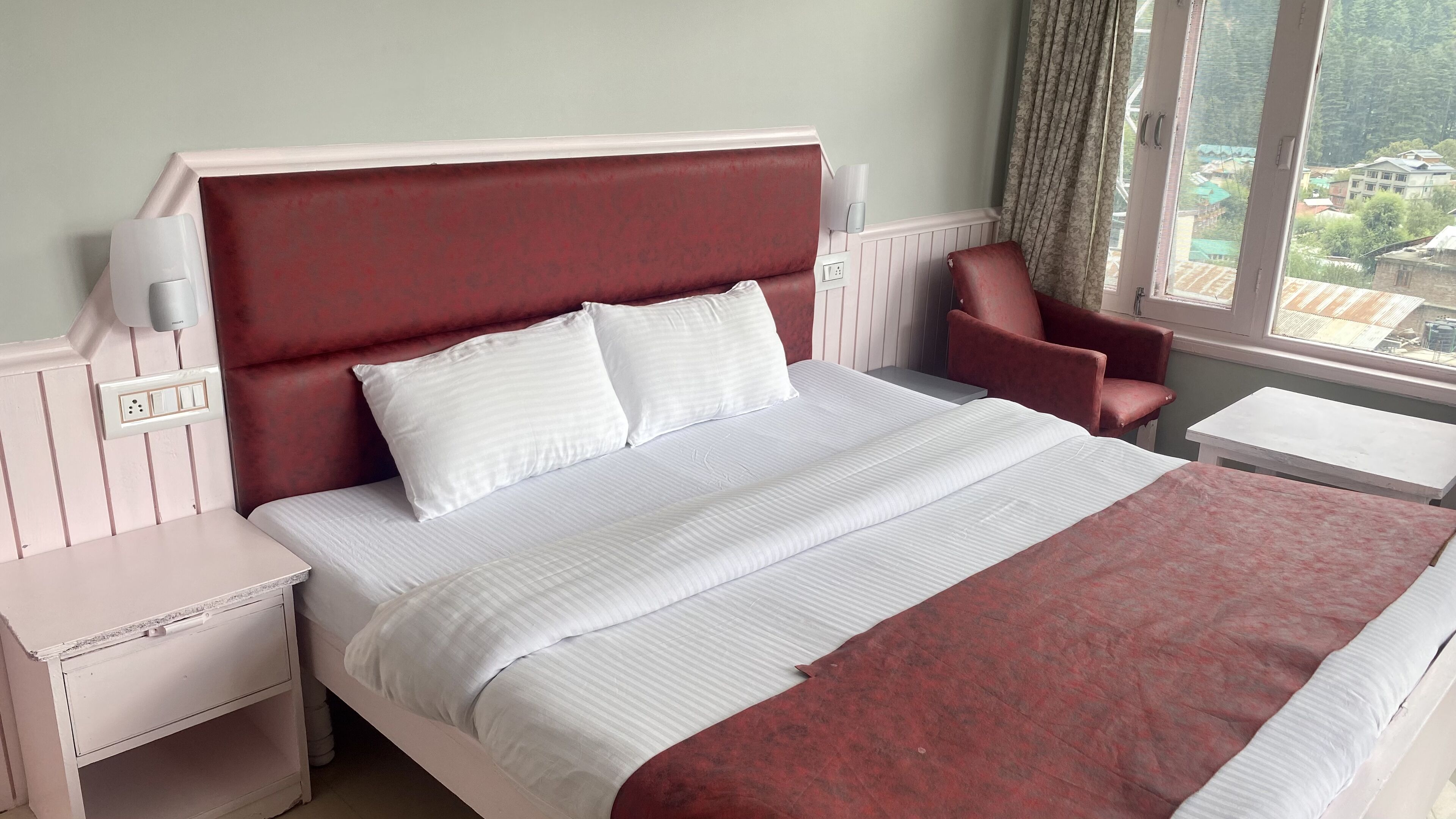 Een net opgemaakt bed met witte lakens en een rood hoofdeinde, een rode fauteuil, een klein nachtkastje en een raam met uitzicht op gebouwen.