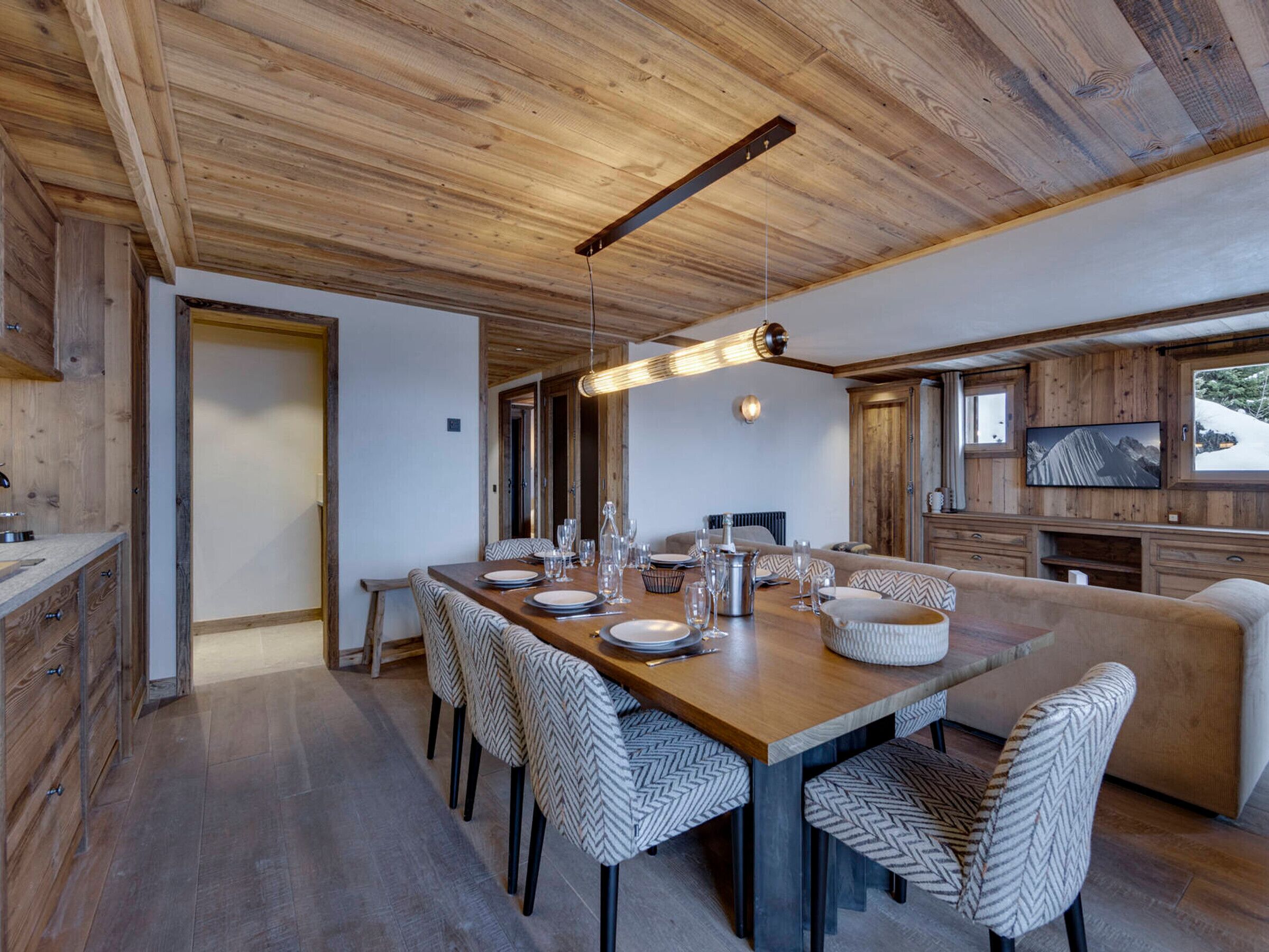 A wooden dining area with a long table, chairs, and a television on the wall.