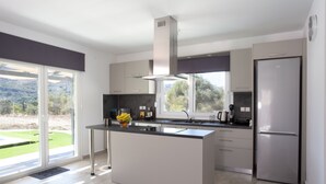 A modern kitchen with a central island, stainless steel appliances, and large windows offering a view of the outdoors. - 3-bed Villa in Palaioloutra With Private Pool (Agios Vasileios)