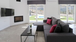 A modern living room with a grey sofa, a flat-screen TV mounted on the wall, and large windows offering a view of a green landscape. - 3-bed Villa in Palaioloutra With Private Pool (Agios Vasileios)