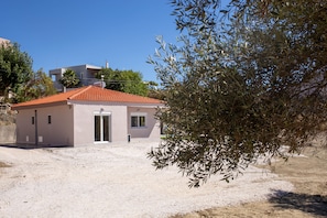 A small building with a red-tiled roof and a white door, surrounded by gravel and olive trees.