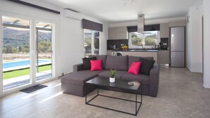 A modern living room with a sectional sofa, a coffee table, and a view of a pool and mountains through large sliding doors. - 3-bed Villa in Palaioloutra With Private Pool (Agios Vasileios)