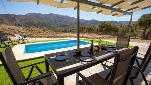 A poolside dining area with a table set for two, overlooking a pool and mountains. - 3-bed Villa in Palaioloutra With Private Pool (Agios Vasileios)