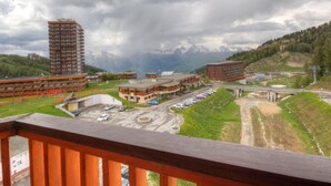 A view of a town with a mix of modern and traditional buildings, a parking area, and a bridge leading to a mountainous area. - Renovated studio, direct access to slopes, balcony, all comfort (La Plagne)