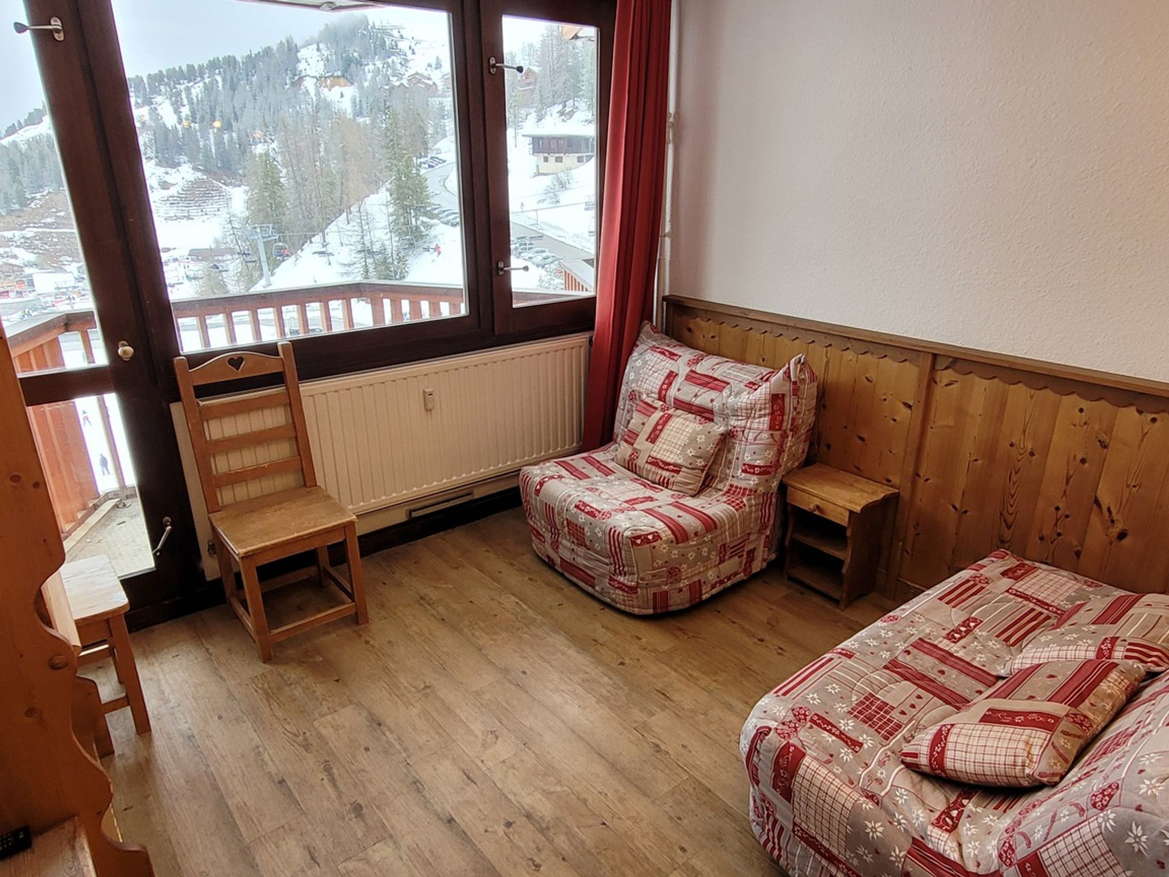 Ein Zimmer mit einer hölzernen Bank, einem kleinen Tisch, einem Bett mit gemusterten Betttextilien und Blick auf eine verschneite Landschaft durch große Fenster.