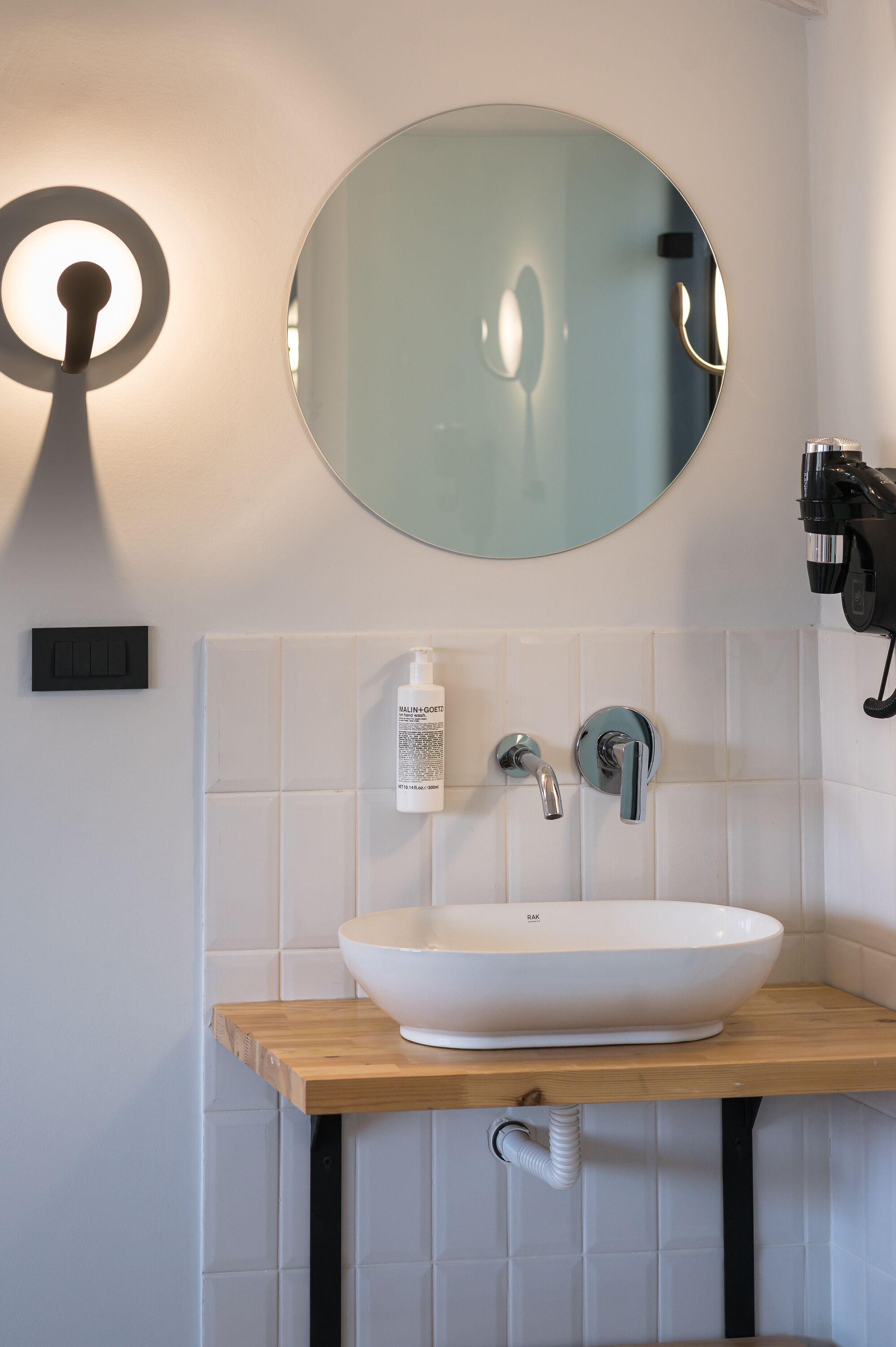 Un baño moderno con un espejo redondo, azulejos blancos, una encimera de madera, un lavabo blanco y un cabezal de ducha de mano.