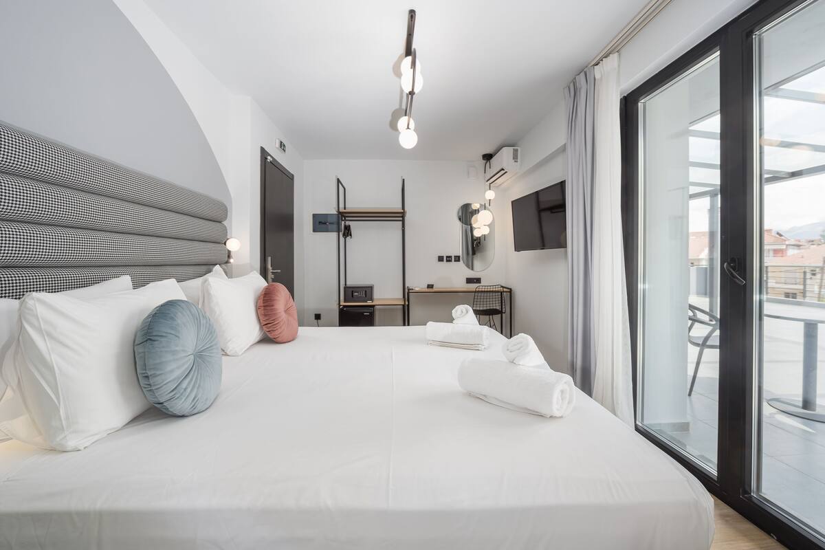 A modern hotel room with a large bed, a desk, a mirror, and a view of the city through large windows.