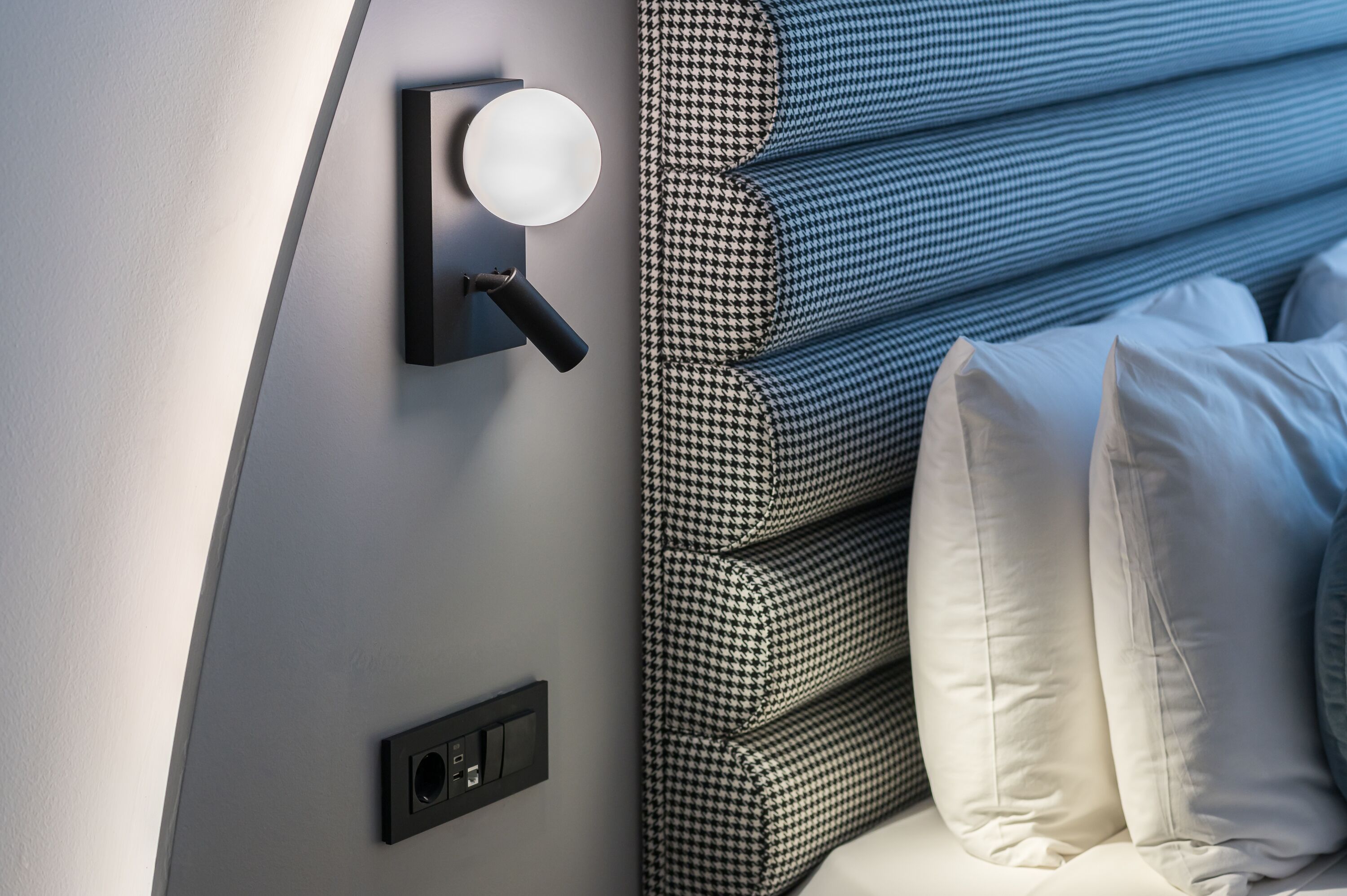 A neatly arranged bed with a houndstooth-patterned headboard, a wall-mounted light fixture, and a power outlet.