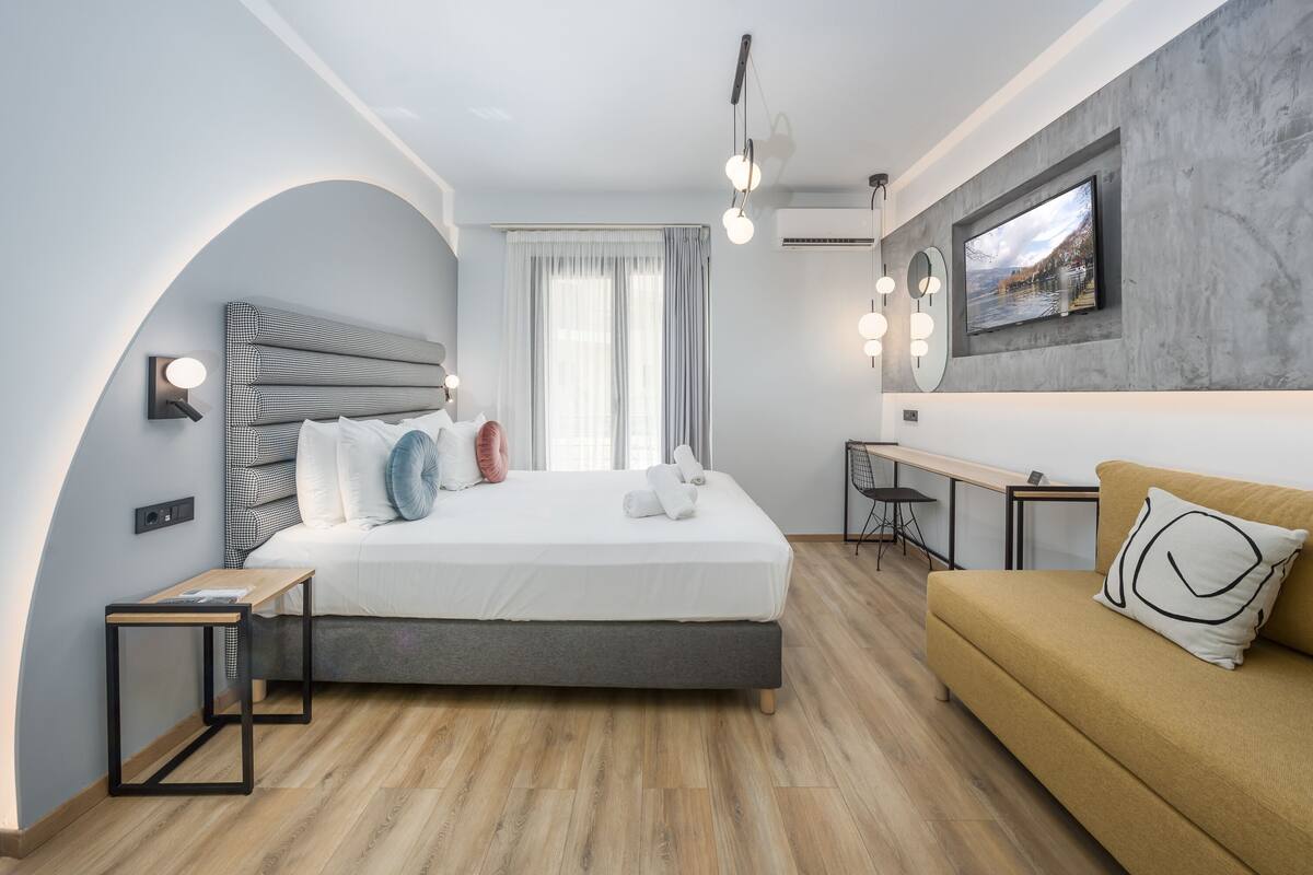 A modern hotel room with a bed, a sofa, a desk, and a TV mounted on the wall.