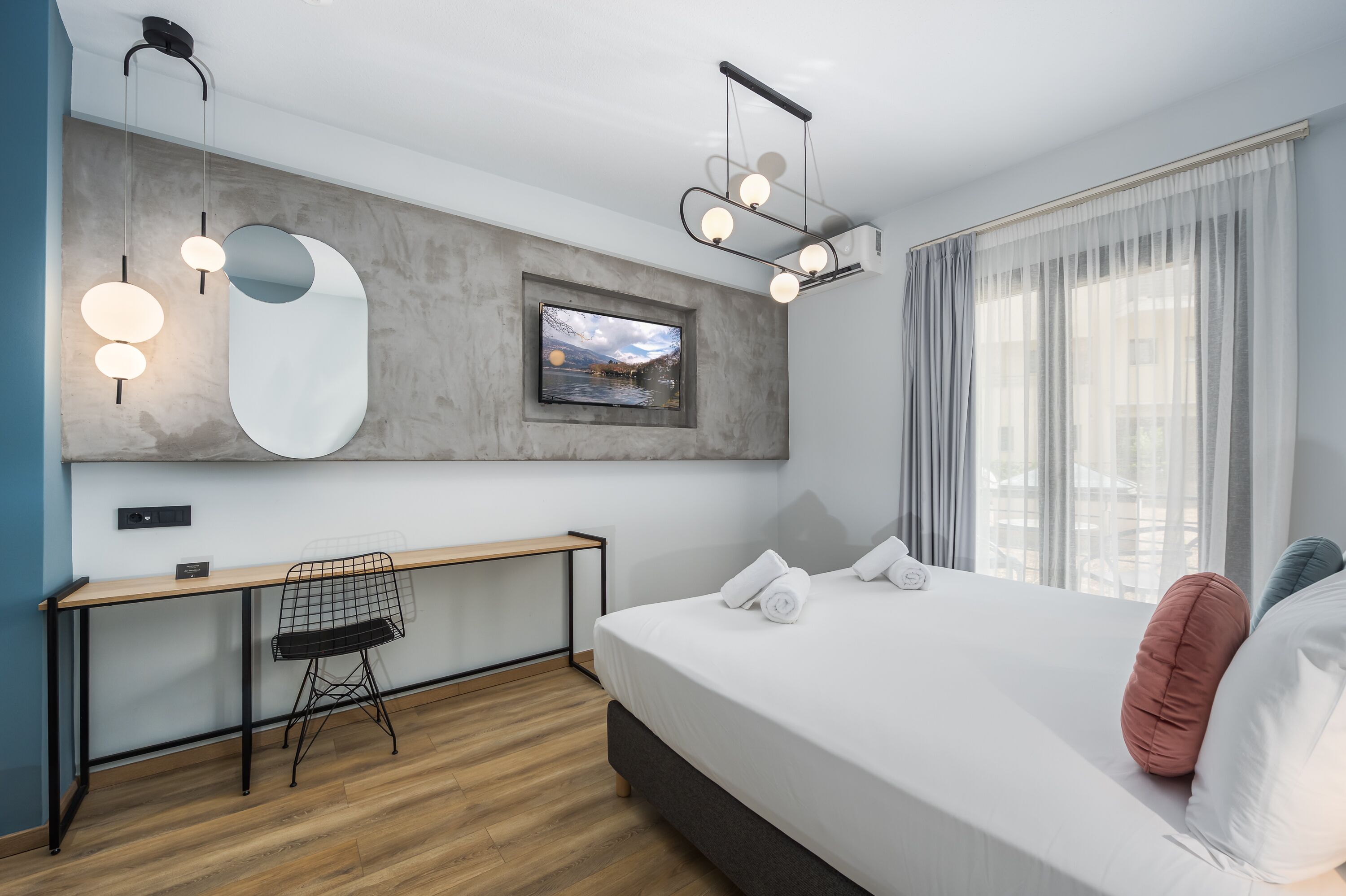 A modern hotel room with a large bed, a desk, and a chair. There is a large window with curtains, a mounted TV, and a ceiling light fixture.