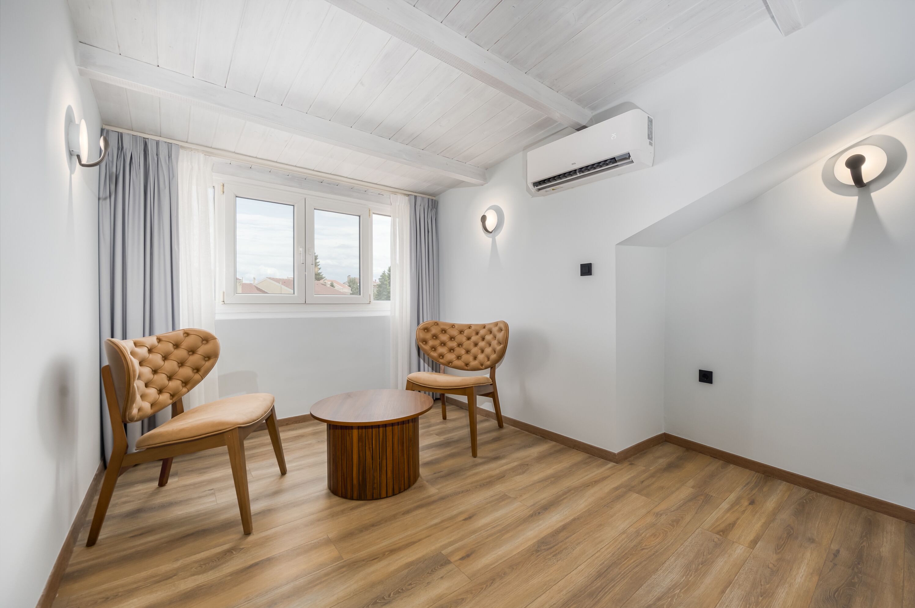 Una habitación con piso de madera, dos sillas, una mesita, una ventana con cortinas y un equipo de aire acondicionado.