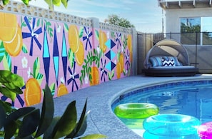 Una piscina con coloridas ilustraciones en la pared, aros hinchables y una zona de descanso con cojines.