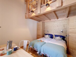 Ein gemütliches Zimmer mit Holzbalkendecke, einem Bett mit blauer Bettwäsche, einem Tisch mit Wasserkocher und Tassen sowie einem Sessel.