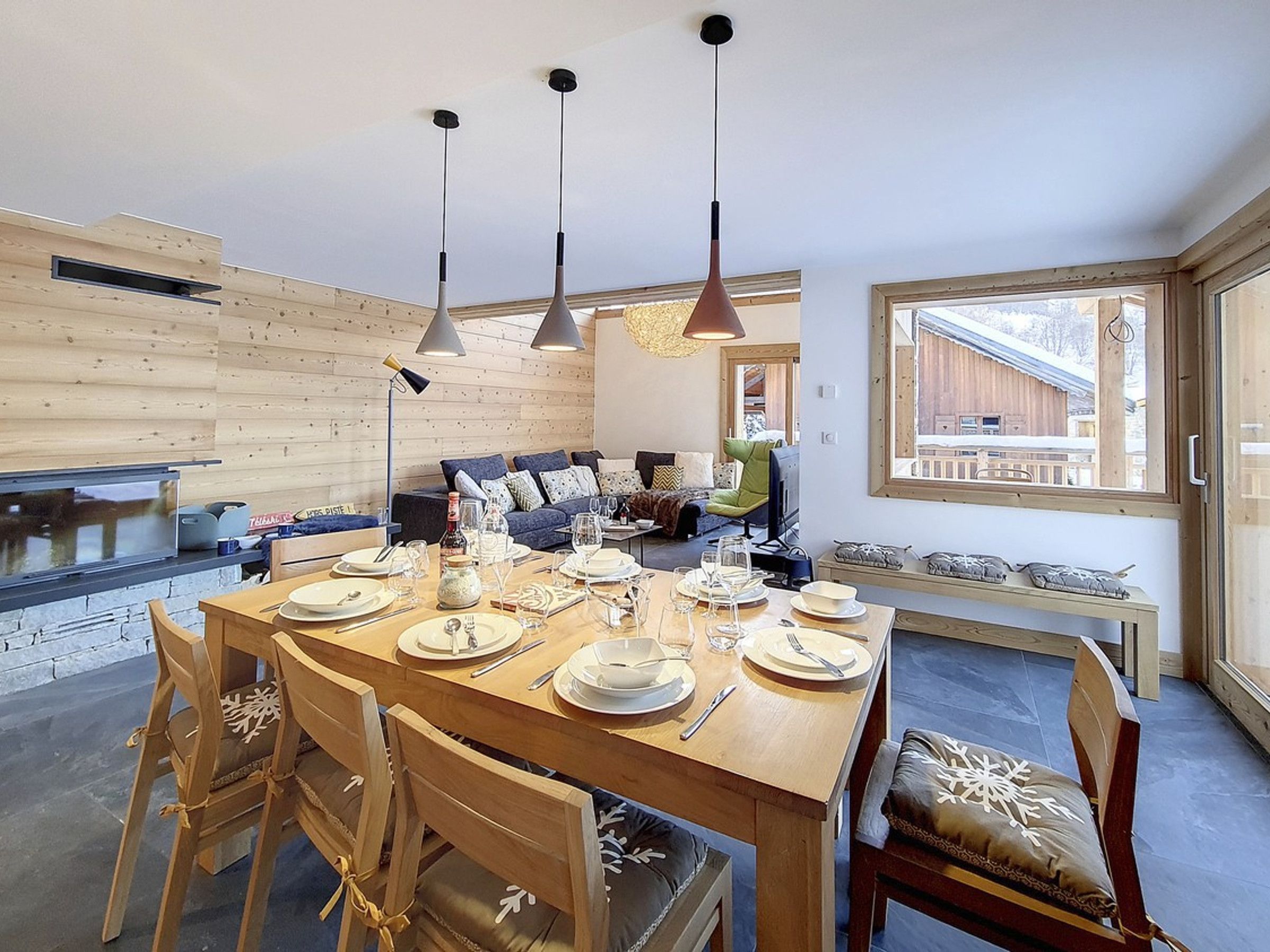A dining area with a wooden table set for four, surrounded by chairs. There are pendant lights above the table, and a window with a view of a wooden building outside.