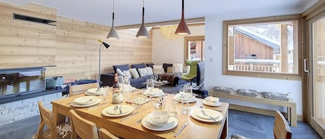 A dining area with a wooden table set for four, surrounded by chairs. There are pendant lights above the table, and a window with a view of a wooden building outside.