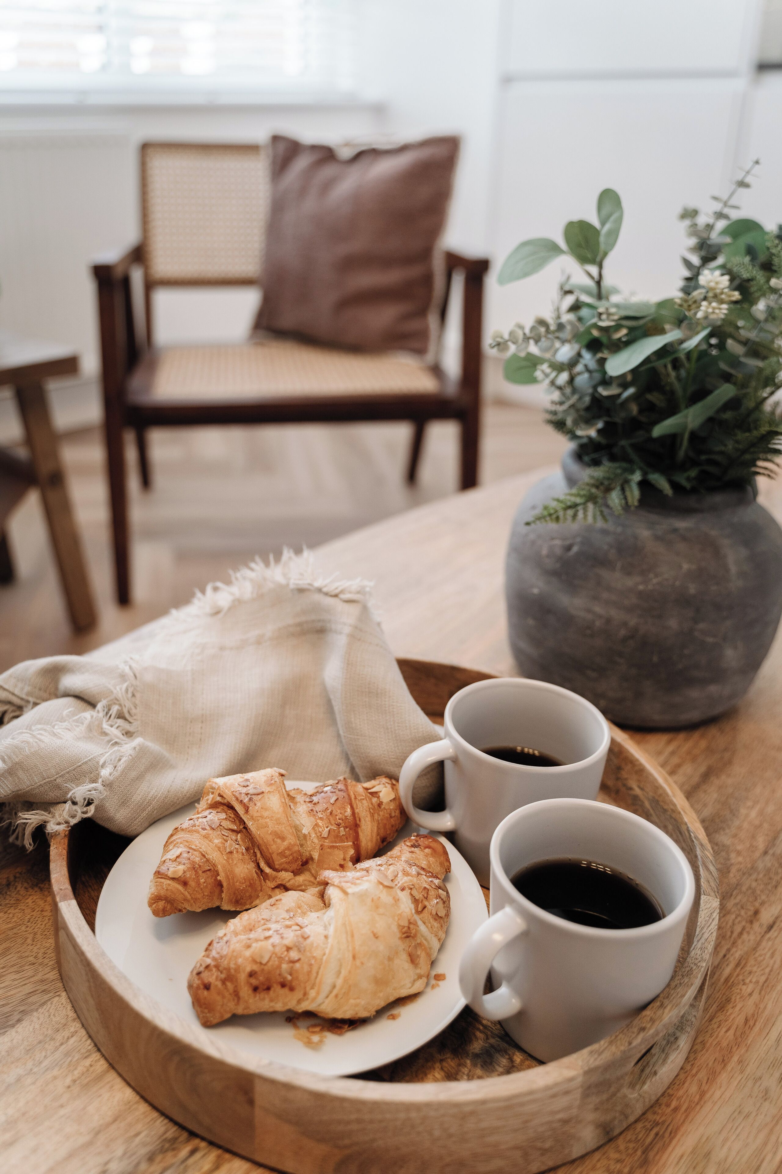Ett träbricka med croissanter och kaffekoppar står på ett bord, bredvid en stol och en krukväxt.