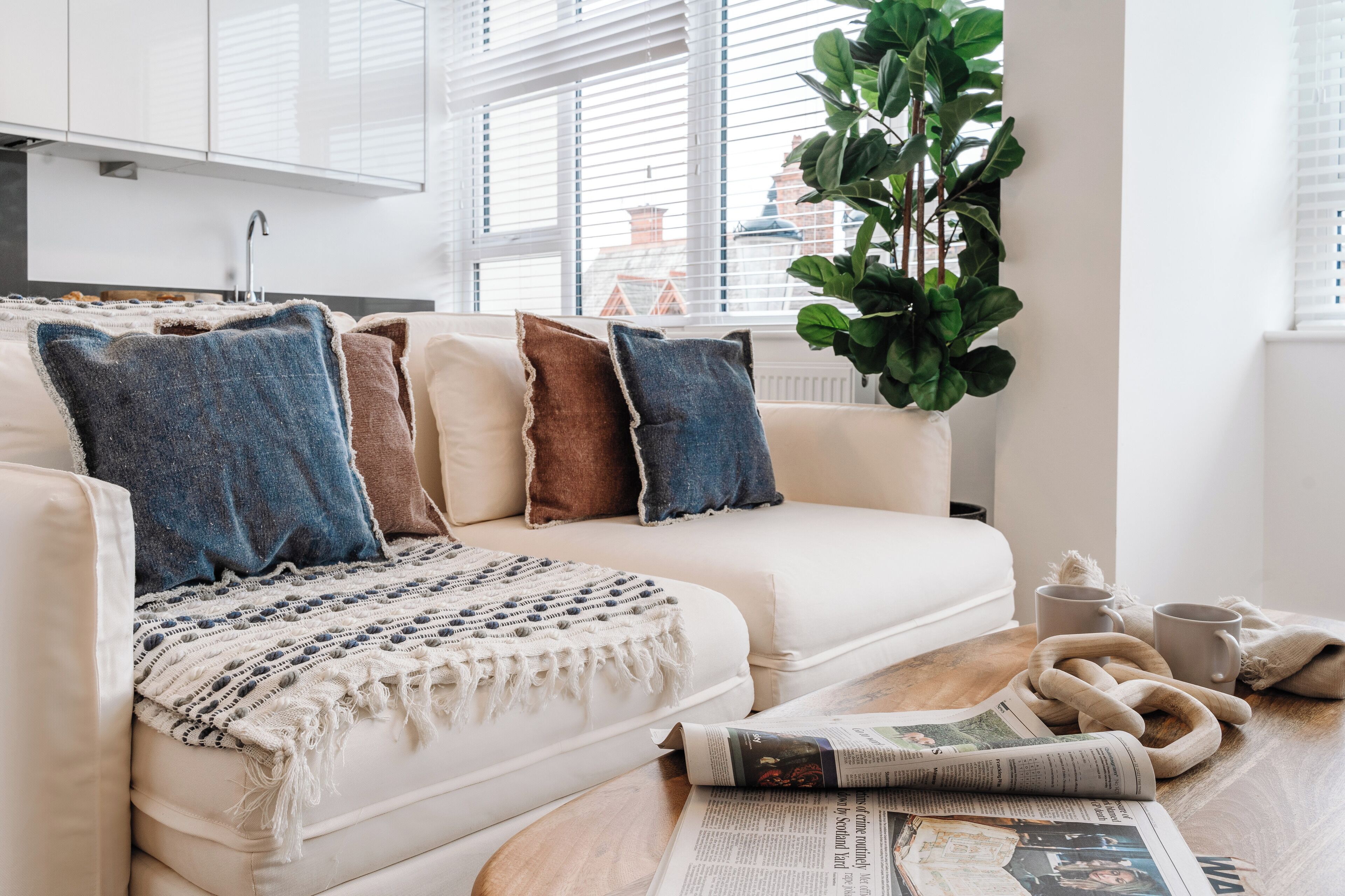 A modern living room with a white sofa, a knitted throw, and a newspaper on a wooden table.