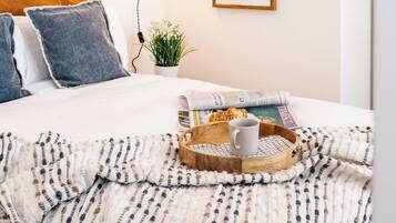 A neatly made bed with a white duvet, a grey pillow, and a blue pillow. There is a tray with a white mug and a plate of food on the bed. A small potted plant is on the nightstand beside the bed.