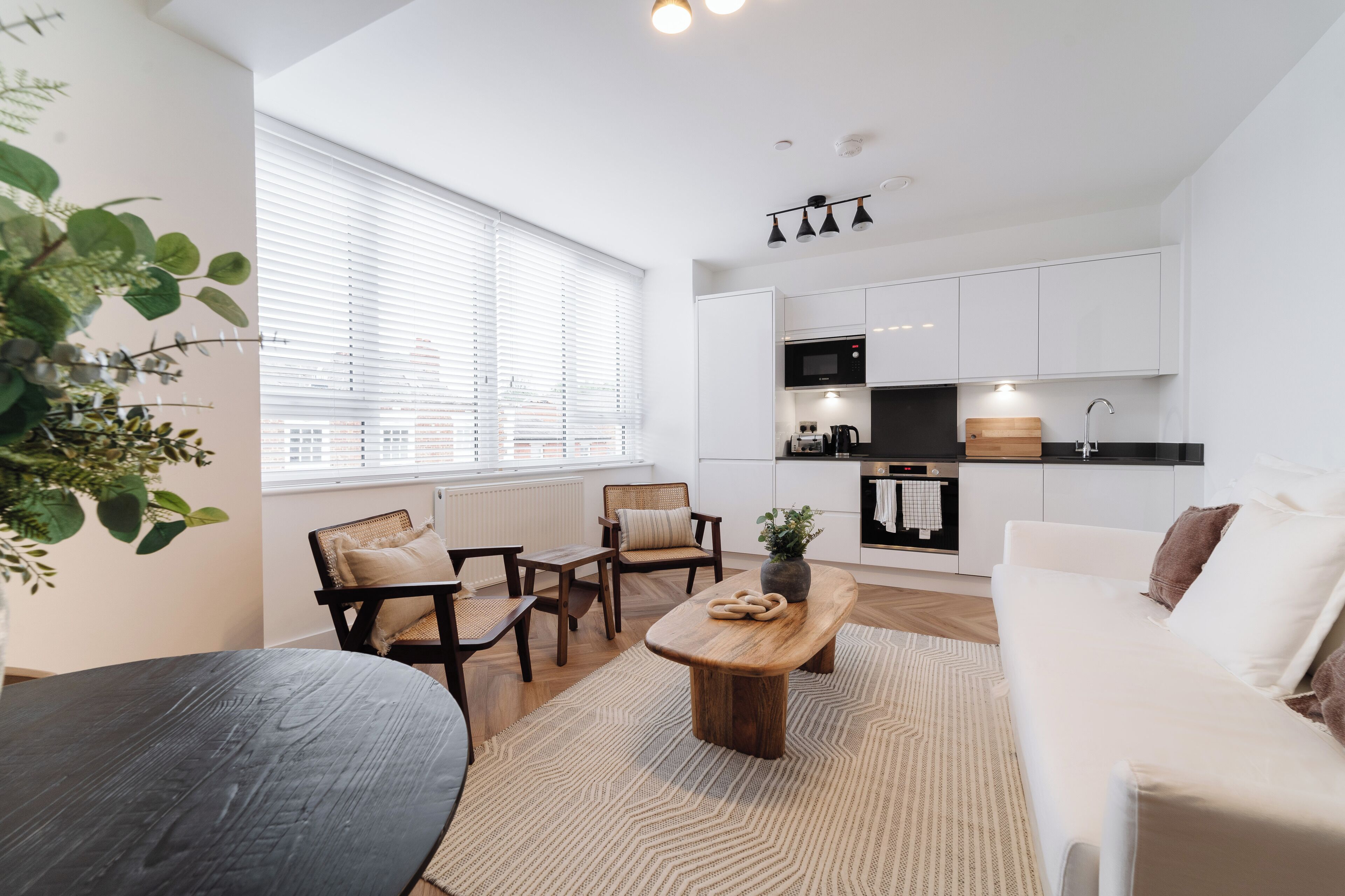 A modern kitchen with white cabinets, a black microwave, and a wooden countertop. There is a dining area with a round table and chairs, and a large window with blinds.
