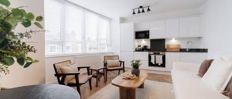 A modern kitchen with white cabinets, a black microwave, and a wooden countertop. There is a dining area with a round table and chairs, and a large window with blinds.