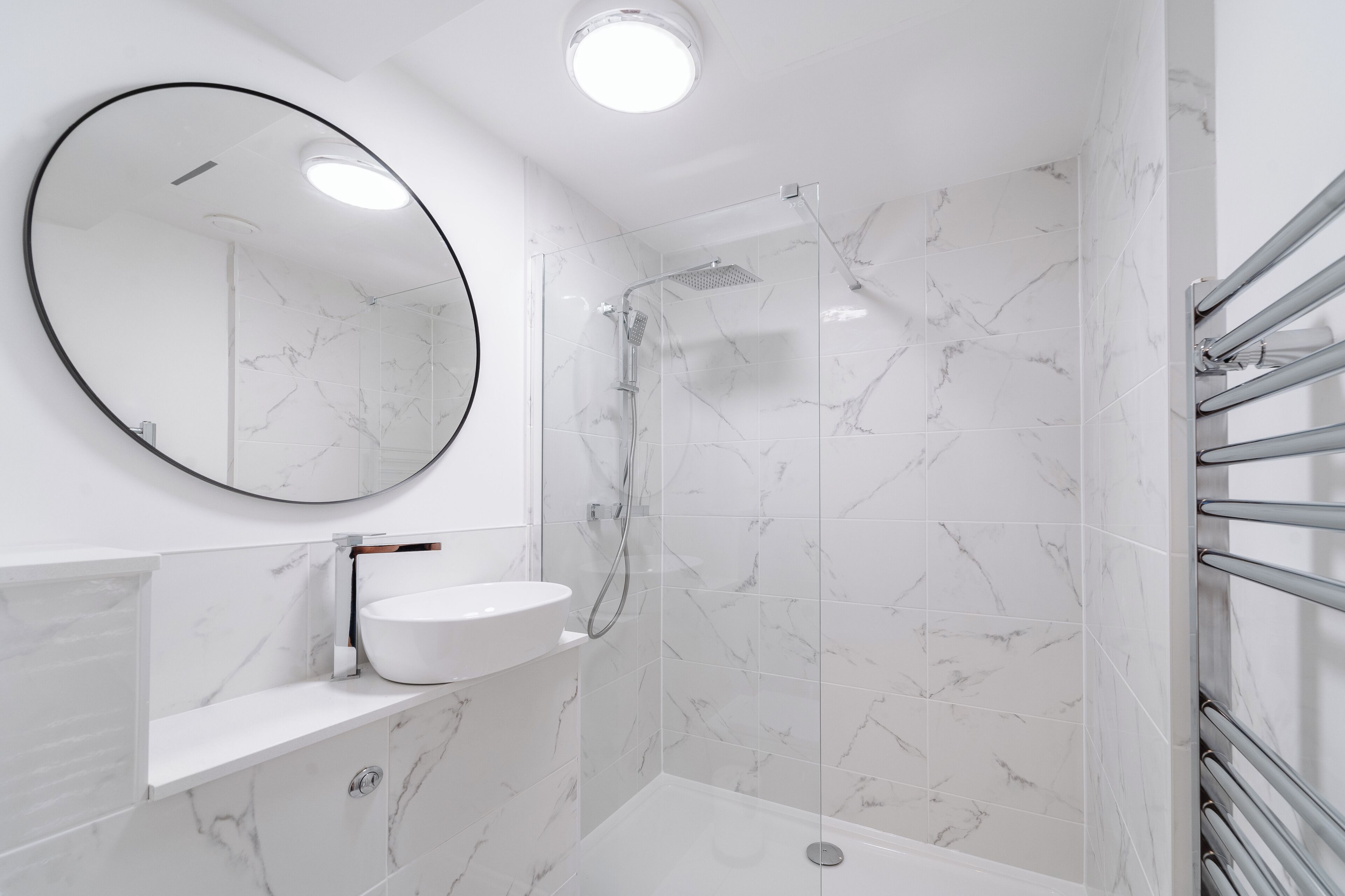 A modern bathroom with a white marble shower, a round mirror, and a white sink.