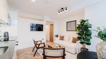 Ein modernes Wohnzimmer mit einem weißen Sofa, einem Holztisch und Sesseln. An der Wand ist ein Fernseher angebracht, es gibt einen Blumentopf und ein großes Fenster.