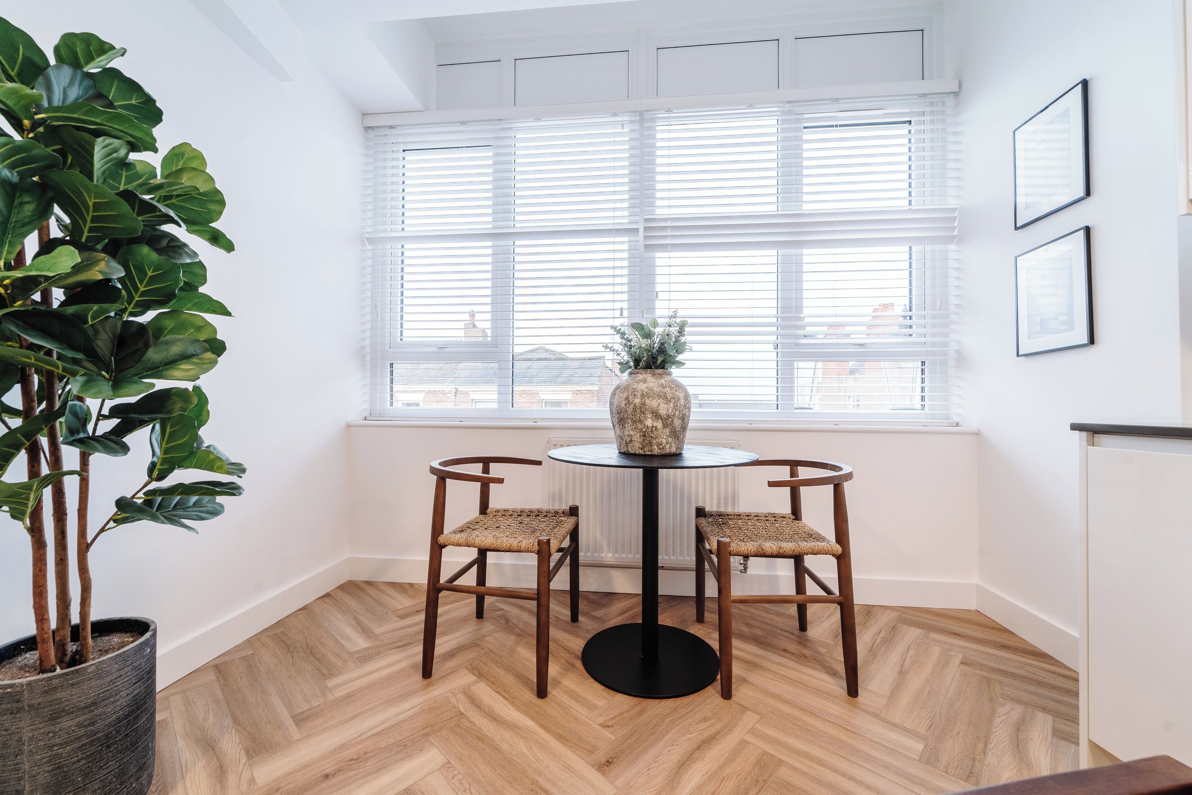 A room with a wooden herringbone floor, a round table with two wicker chairs, a potted plant, and a window with blinds.