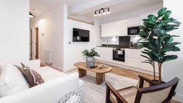 Ein modernes Wohnzimmer mit einer Couch, einem Couchtisch mit einer Vase, einer großen Pflanze und einer Küchenzeile mit weißen Schränken und Geräten.
