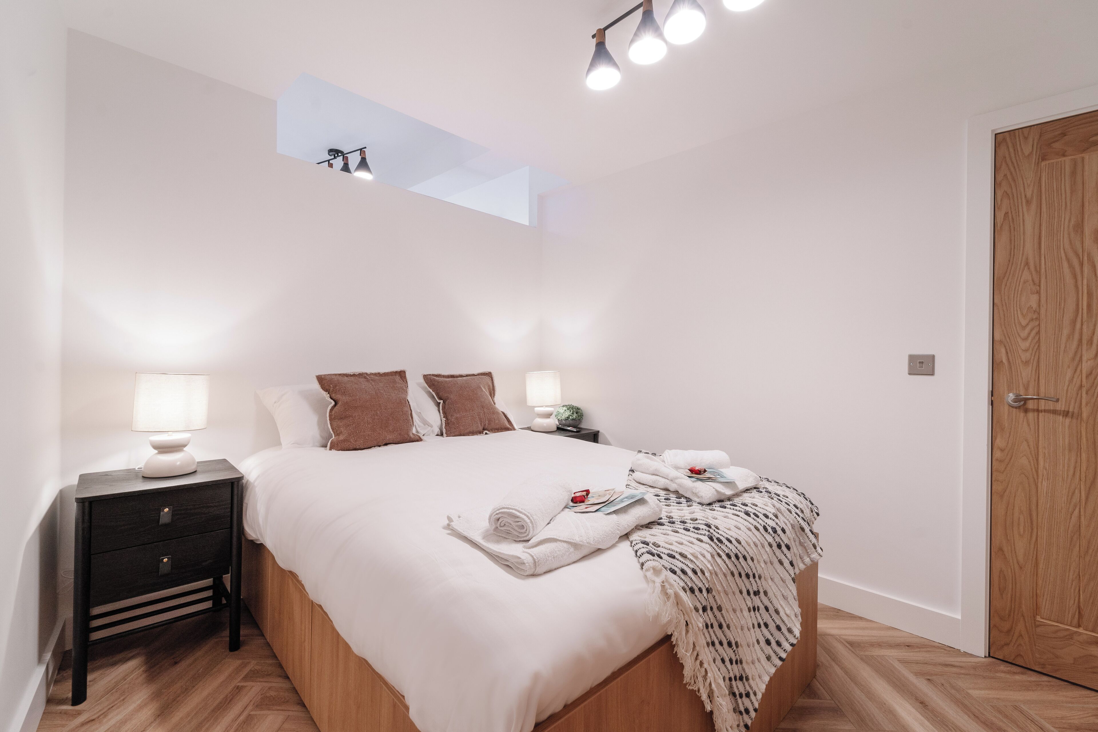 Ein Schlafzimmer mit einem Bett, Nachttischen, einer Stehlampe, einem kleinen Tisch mit Lampe, einer Holztür und einem Oberlicht.