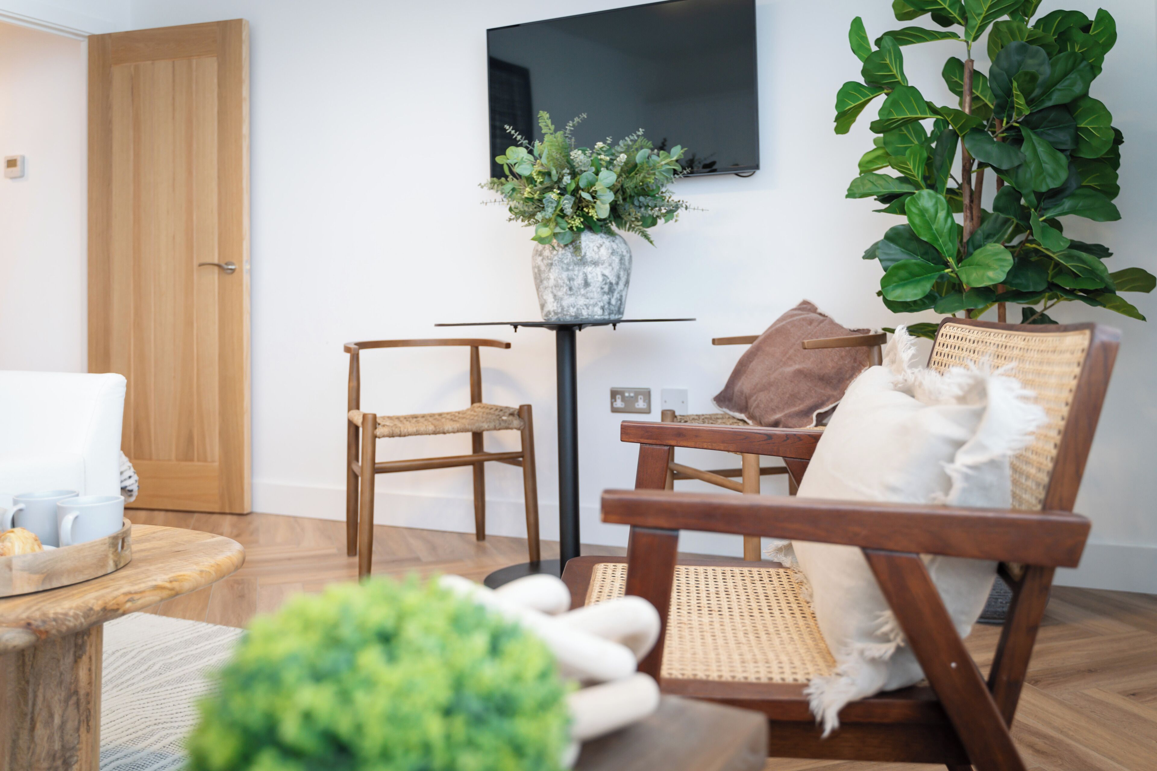 A cozy living room with a wooden floor, a wicker chair, a small table with a vase of plants, and a television mounted on the wall.