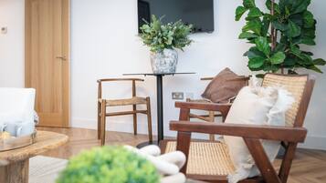 Un salón acogedor con piso de madera, un sillón de mimbre, una mesita con un jarrón de plantas y un televisor instalado en la pared.