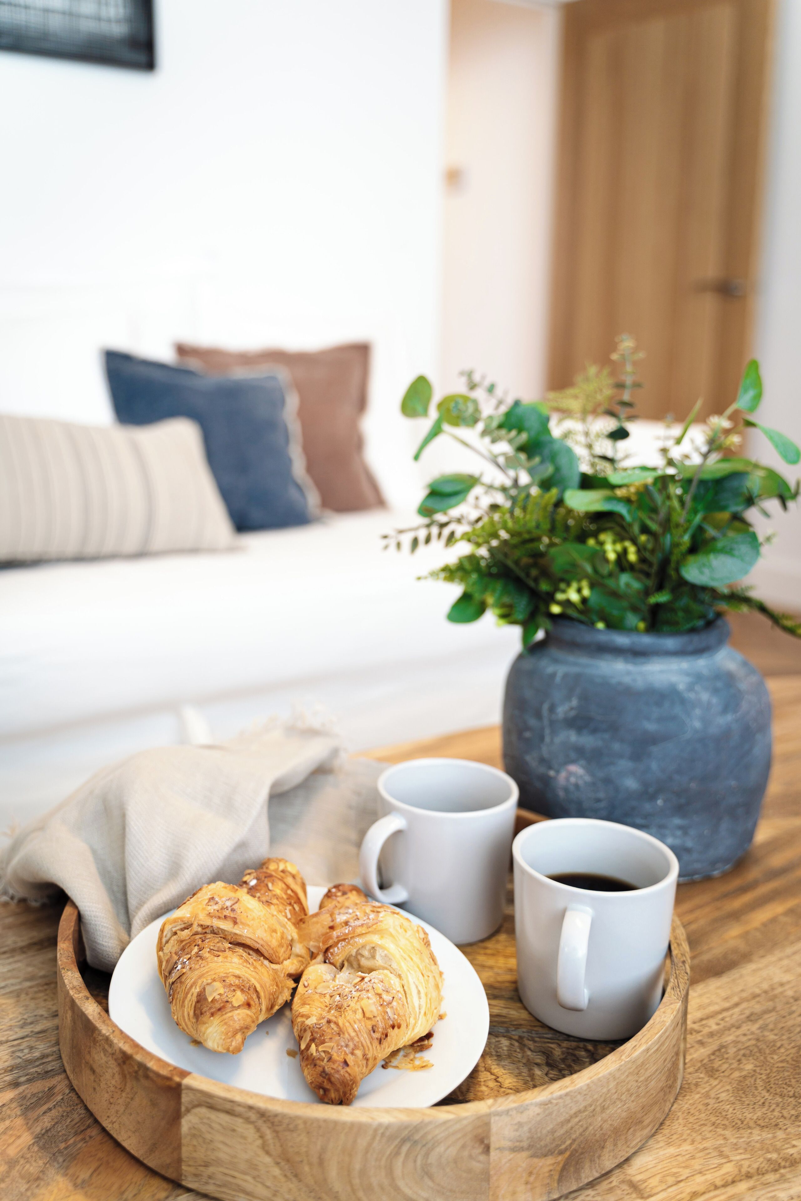 A wooden tray with croissants, two white coffee cups, and a vase with green plants.