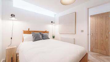 A bedroom with a large bed, two bedside lamps, a small bedside table, a wooden headboard, a radiator, and a wooden door.