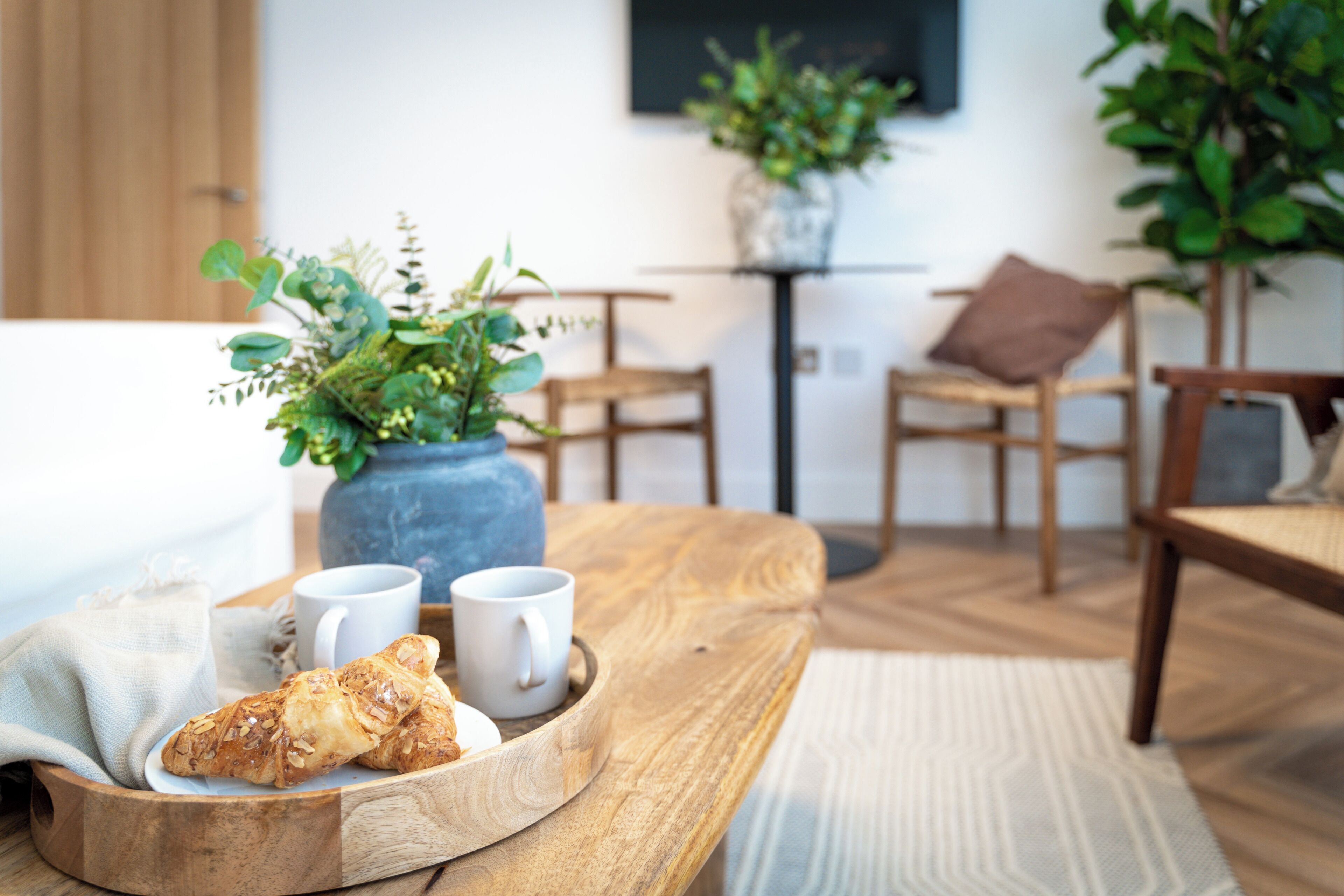 Ein gemütliches Zimmer mit einem Holztisch, einer Platte mit Croissants und Kaffeetassen sowie einer Topfpflanze.