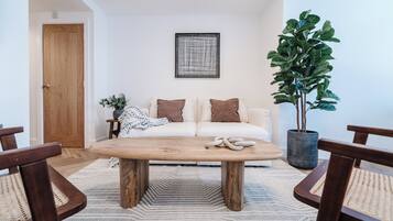 A living room with a wooden coffee table, a white sofa with brown cushions, a potted plant, and a framed artwork on the wall.