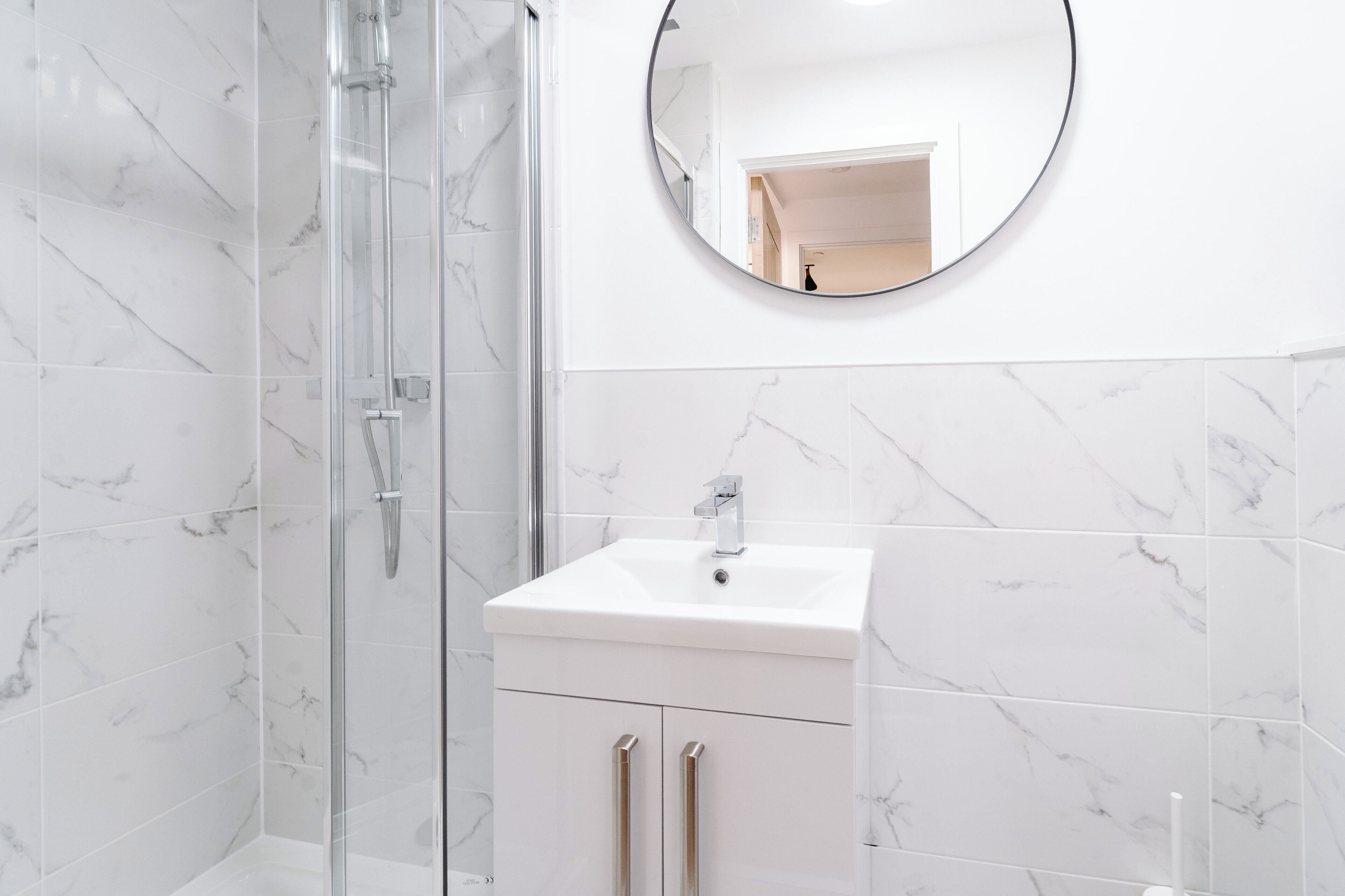 A bathroom with a glass shower enclosure, a white sink, and a round mirror.