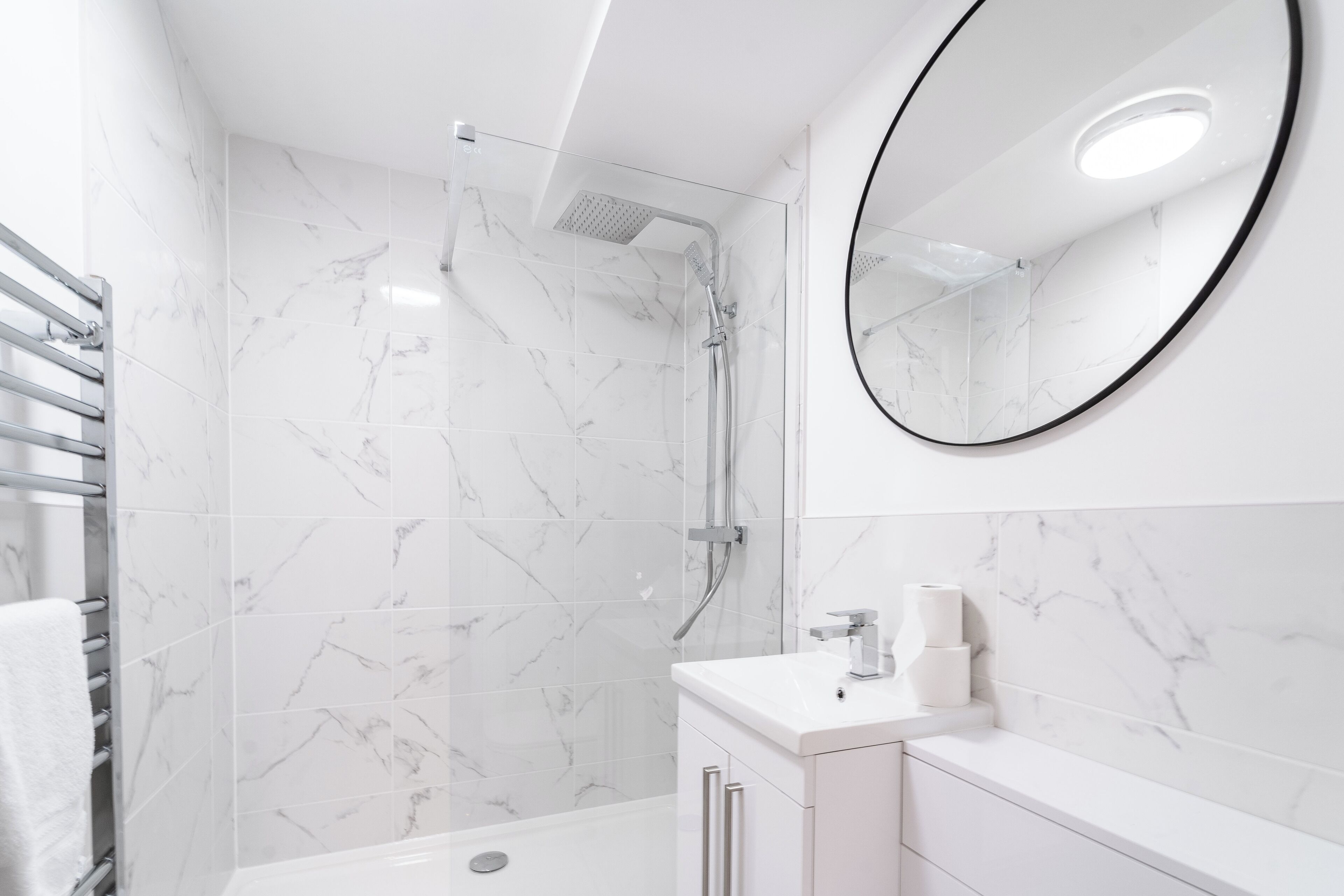 Un baño moderno con ducha envuelta en vidrio, azulejos de mármol blanco, un espejo redondo y un lavamanos blanco con encimera.