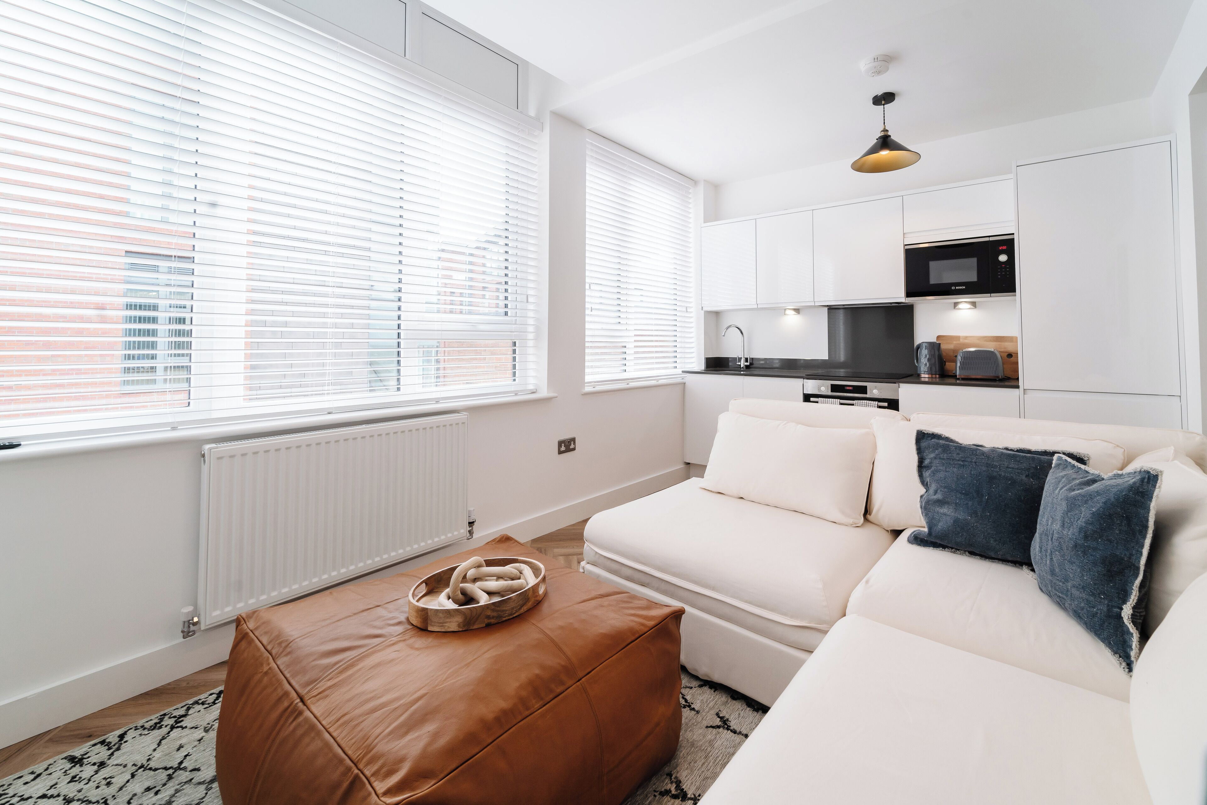Una sala de estar moderna con un sofá blanco, un otoman marrón y una cocina con gabinetes y electrodomésticos blancos.