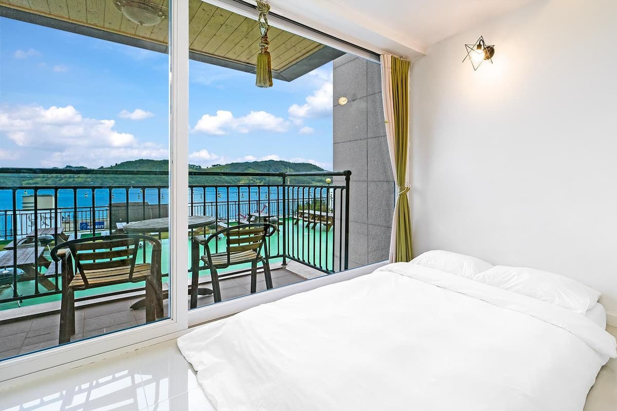 Une chambre dotée d’une grande fenêtre donnant sur un balcon avec vue sur la mer et les collines.
