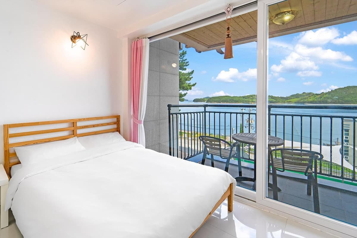Une chambre à coucher avec un lit en bois, une table de chevet blanche et une grande porte-fenêtre coulissante donnant sur un balcon avec vue sur un lac et des collines.