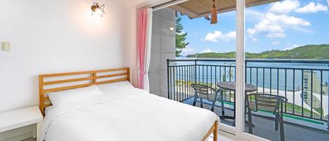 A bedroom with a wooden bed, a white nightstand, and a large sliding glass door leading to a balcony with a view of a lake and hills.