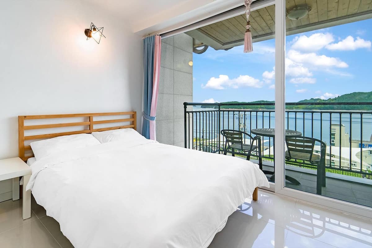 Une chambre avec un lit, un banc, une vue sur la mer et un balcon avec des chaises.