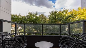 Ein Balkon mit zwei schwarzen Korbstühlen und einem kleinen runden Tisch, mit Blick auf einen Garten mit Bäumen und einem Gebäude im Hintergrund.