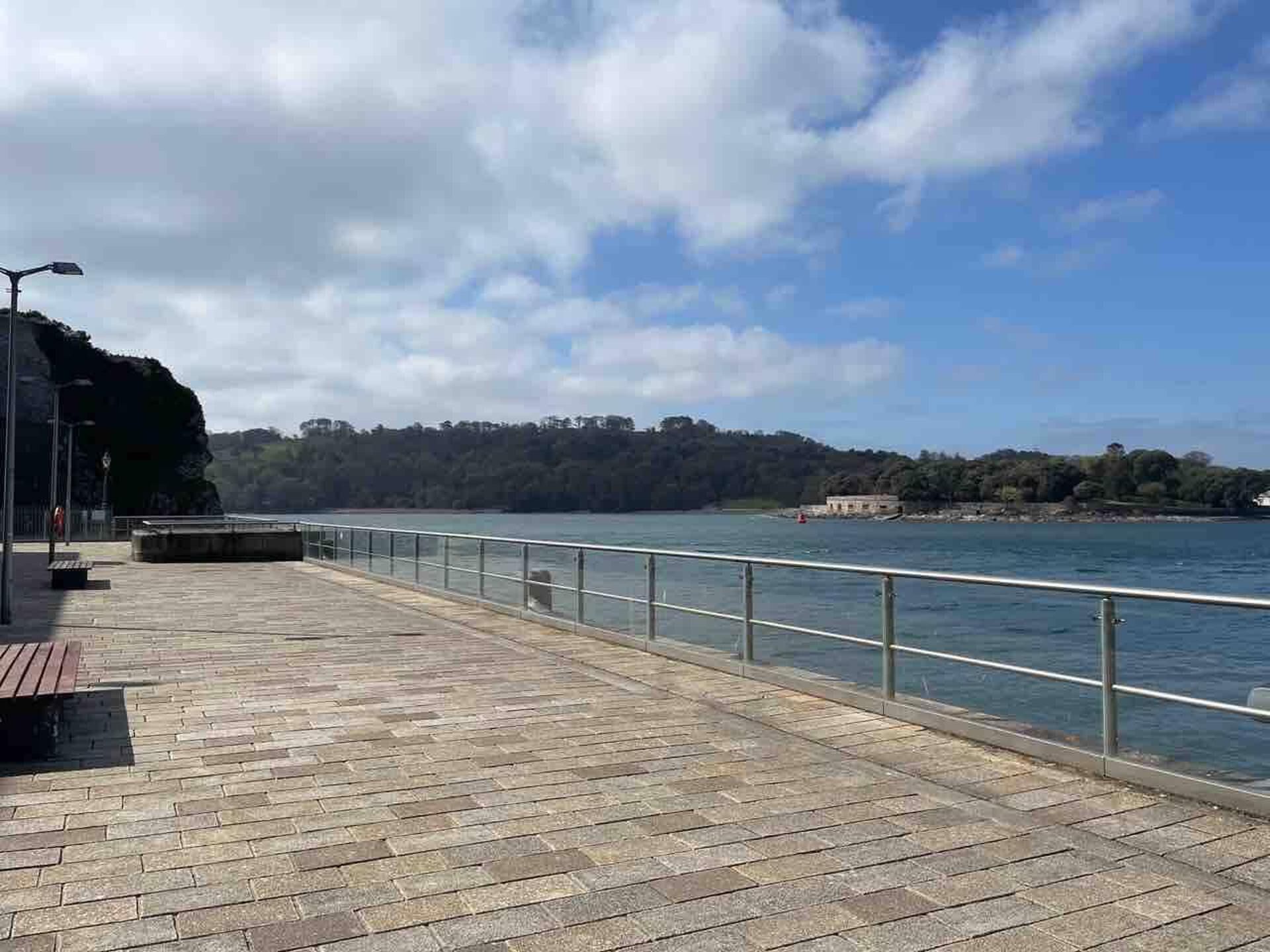 Un paseo marítimo con un camino pavimentado, barandilla metálica y bancos.